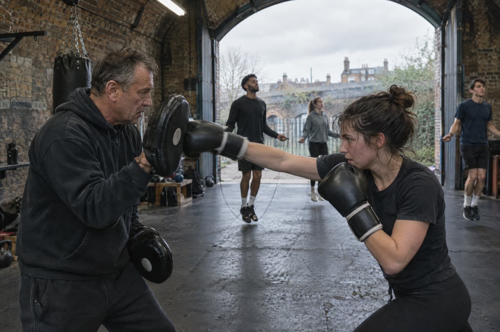 A coach holding pads for a member in a railway-arch boxing gym, others skipping behind.
