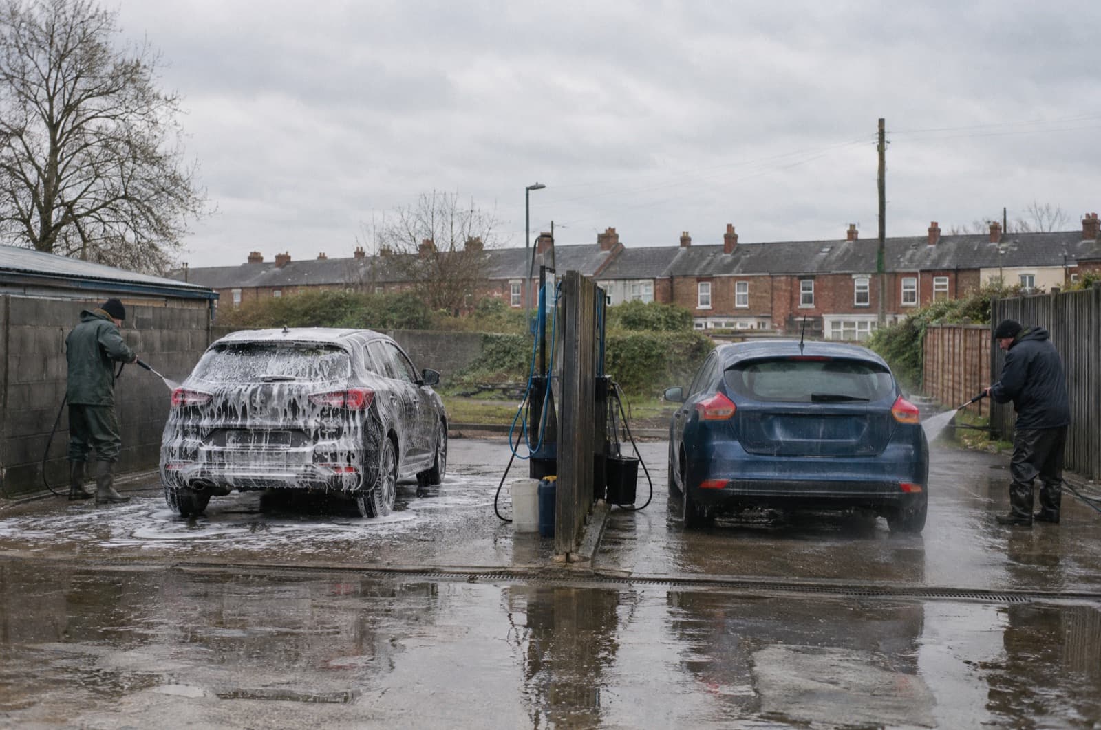 Two cars being washed side by side on a forecourt under a grey sky.