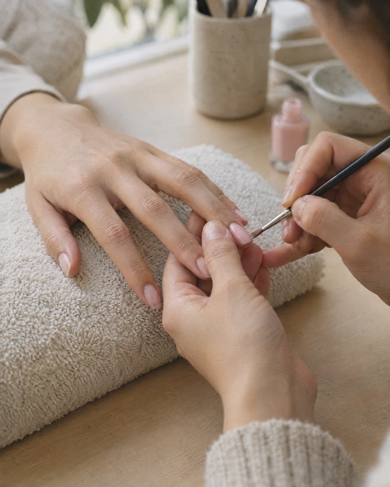 A single nail being painted a milky pink, the client’s hand resting on a cream towel.