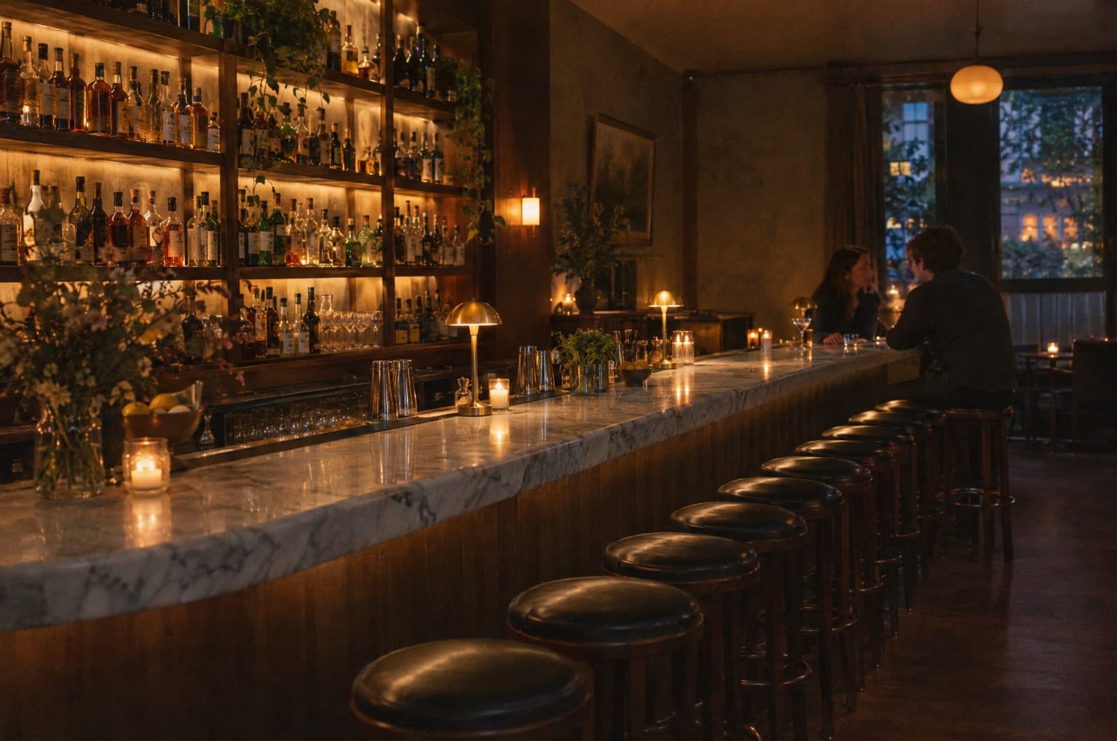 A small cocktail bar early in the evening, a dozen stools along the marble and two people talking at the end.