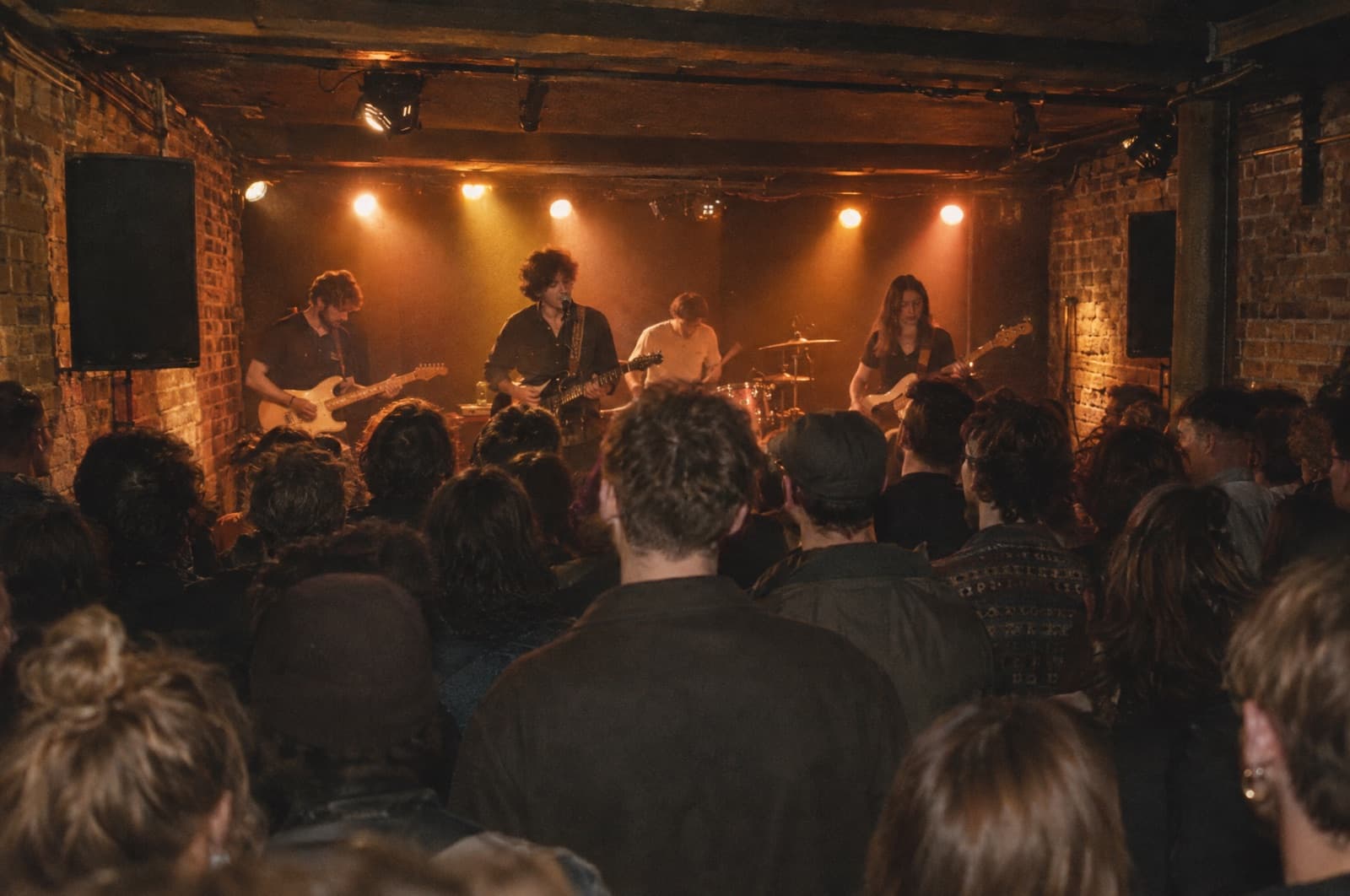 A four-piece band on a low stage in a small brick-walled venue, the room three-quarters full, seen from the back over the crowd.