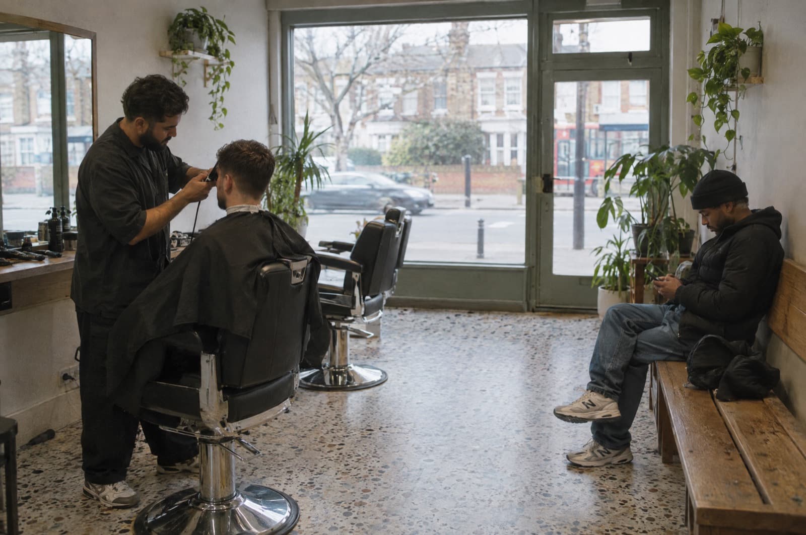 A two-chair barbershop on a weekday afternoon, one customer in the chair and one waiting on the bench.