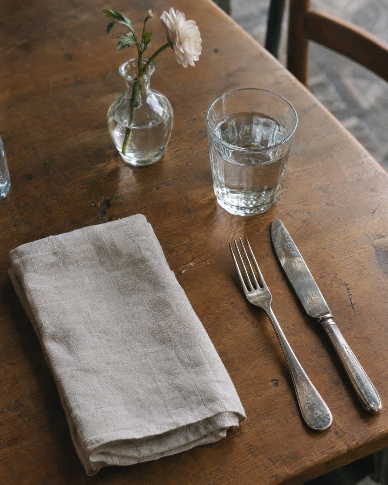 A laid table: linen napkin, worn cutlery, a water glass and a single stem in a small vase.